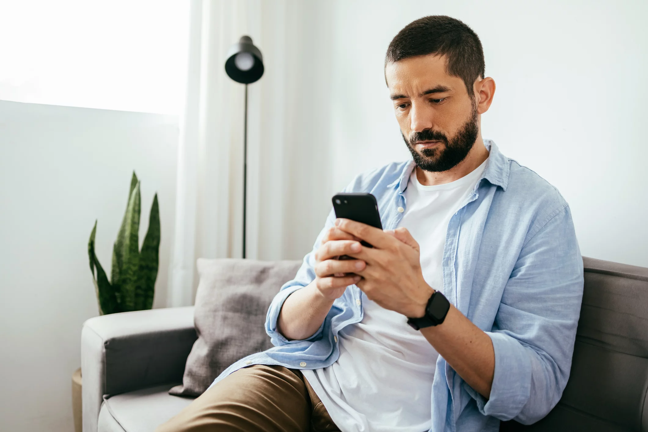 Young brazilian man sitting on sofa using smartphone