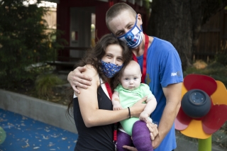 Two parents with masks holding their baby who has both legs in casts