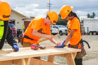 Three people working on constructing a picnic table with one person hammering a nail into the top of it