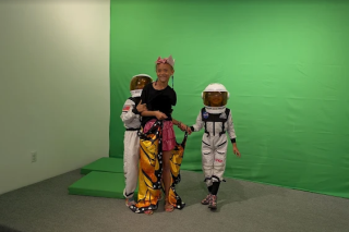 Three children, two dressed as astronauts and one dressed as a butterfly, standing in front of a greenscreen