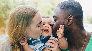 Thumbnail photo of two parents kissing their baby